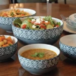 A beautifully styled wooden dining table featuring a set of grey and white patterned ceramic serving bowls filled with fresh salad, soup, pasta, and roasted vegetables.