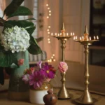 A warm, festive home decor setting featuring two tall, lit traditional brass oil lamps (diyas) on a table. The scene is decorated with fresh white hydrangeas and pink cosmos flowers in vases, with soft string lights glowing in the background.