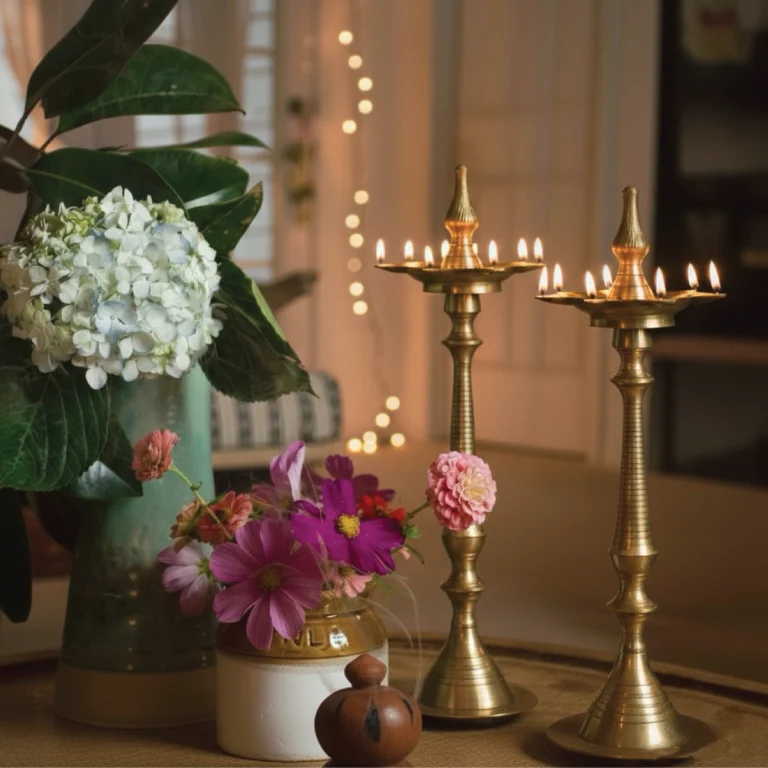 A warm, festive home decor setting featuring two tall, lit traditional brass oil lamps (diyas) on a table. The scene is decorated with fresh white hydrangeas and pink cosmos flowers in vases, with soft string lights glowing in the background.