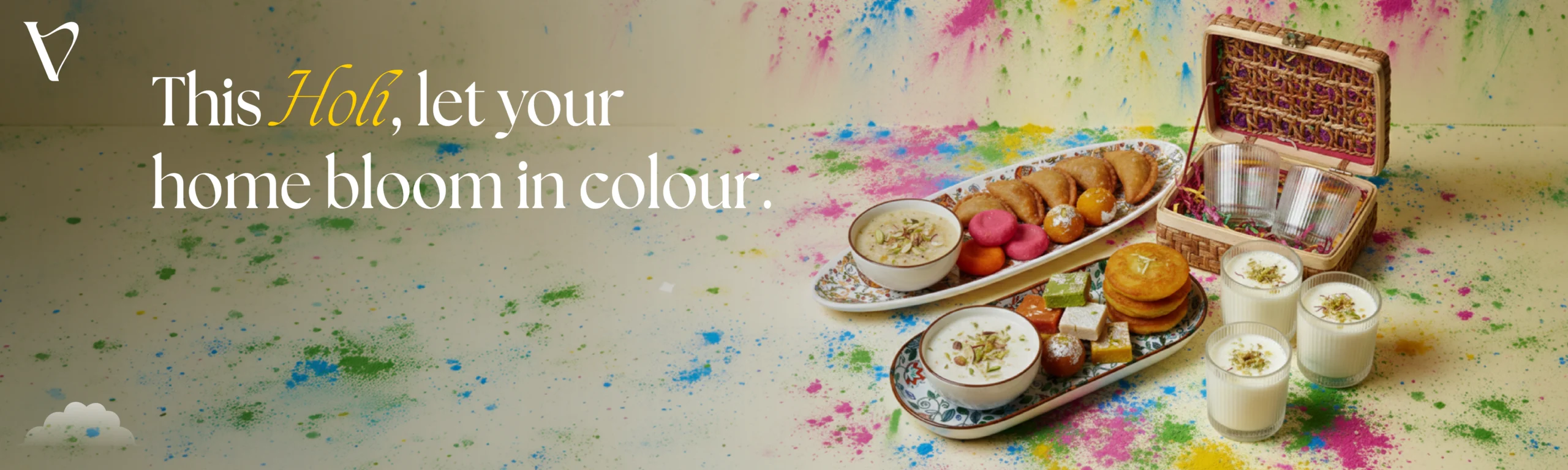 Holi festive promotional banner reading "This Holi, let your home bloom in colour." The image features a vibrant spread of traditional Indian sweets, gujiya, and thandai served in patterned ceramic platters and ribbed glass tumblers, surrounded by colorful gulal powder splashes.